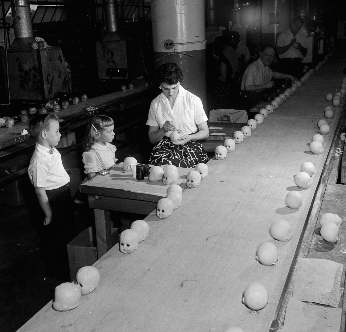 circa 1955: A young boy and girl watching workers putting eyes into dolls' heads as they pass along a conveyor belt at the Ideal Toy Company in Jamaica, Long Island, USA. The company is one of the largest toy manufacturers in the world. (Photo by Orlando /Three Lions/Getty Images)
